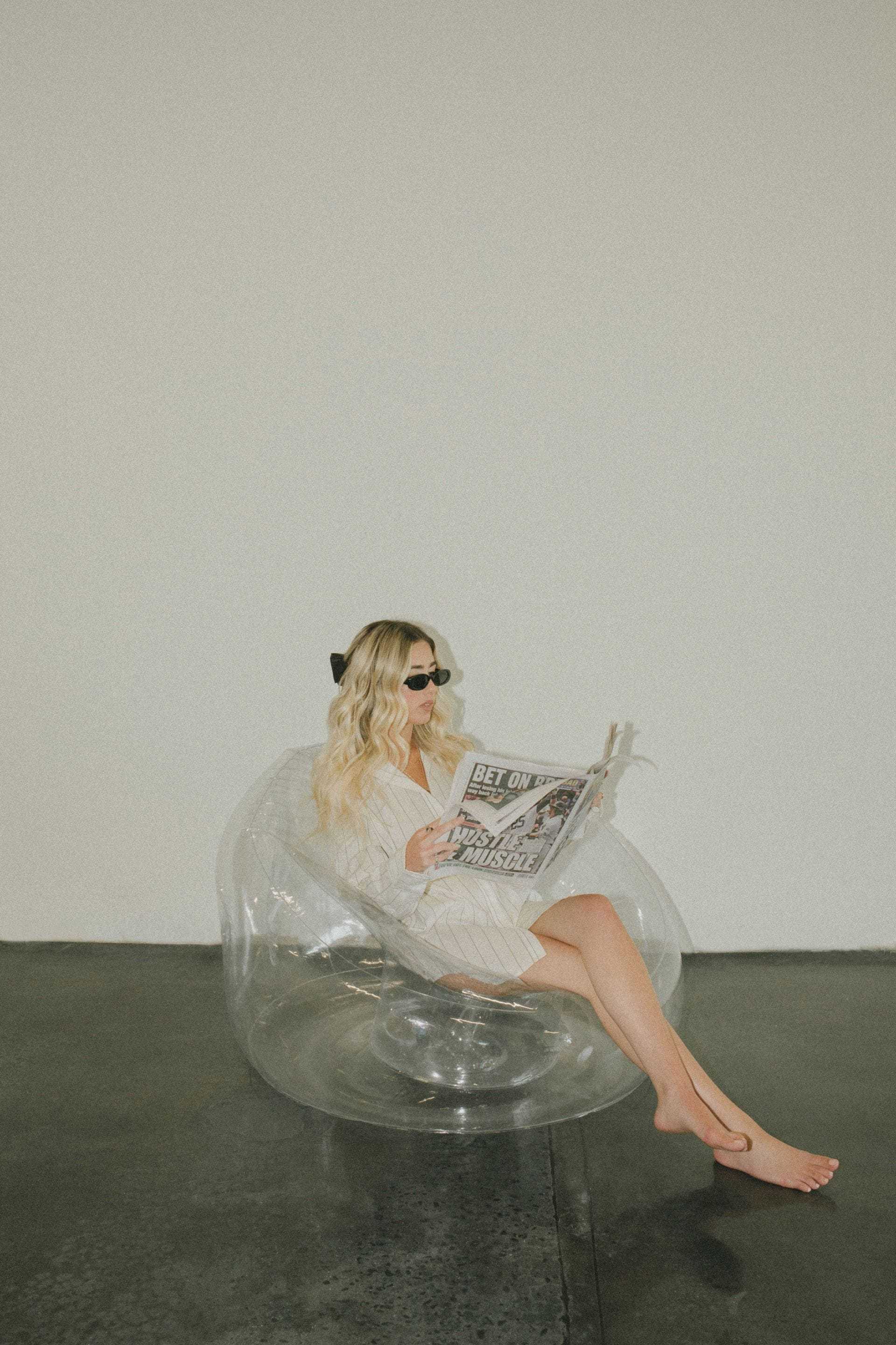 Woman in a blazer reading a newspaper on a transparent chair against a plain wall.