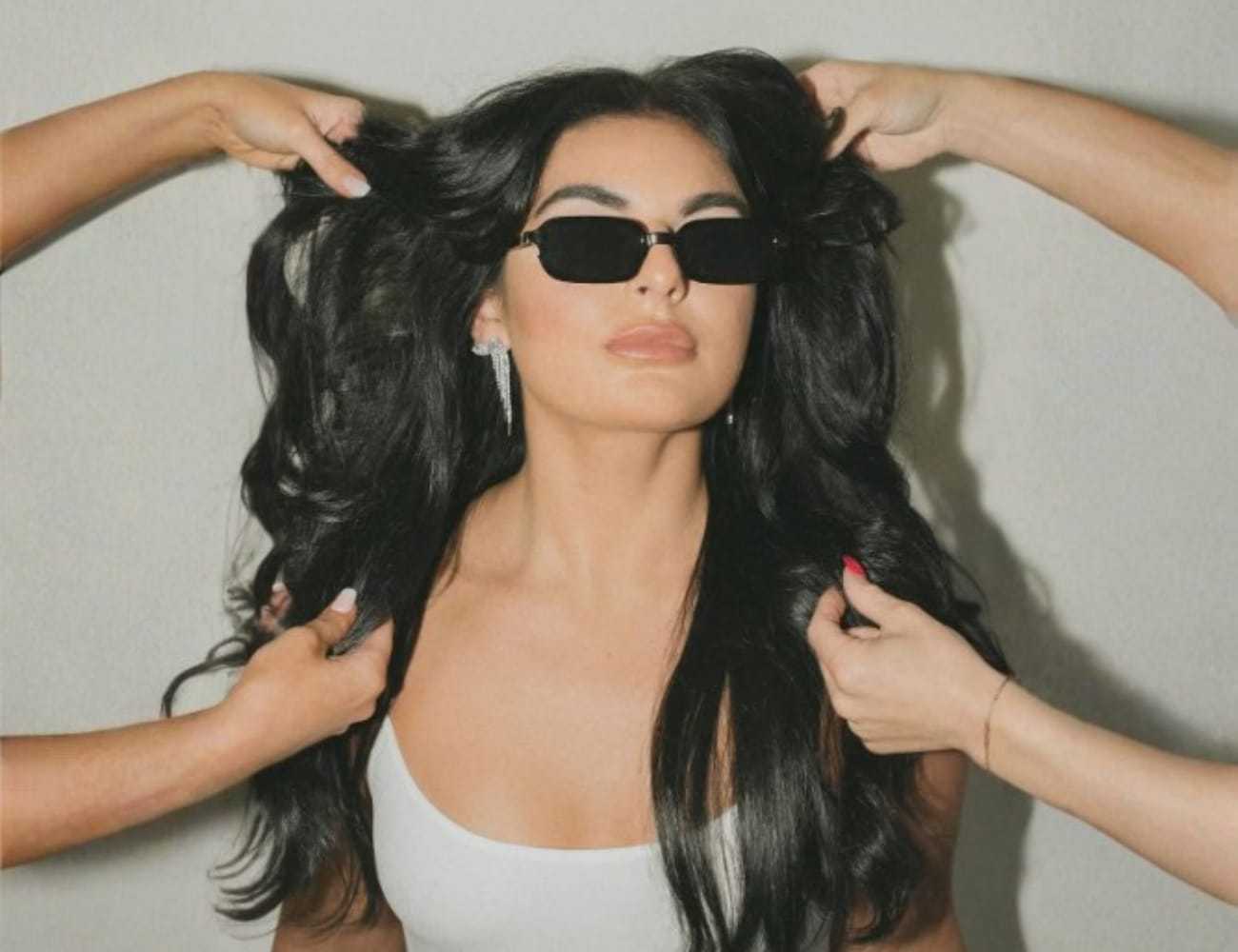 Woman with long hair styled by two people, wearing sunglasses and a white top.