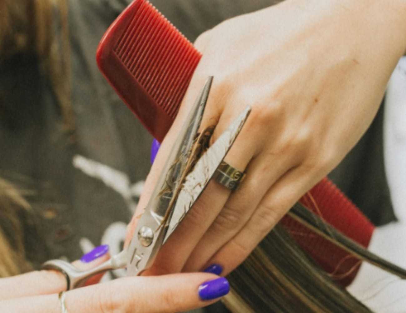 Cutting hair with scissors and a red comb, close-up of stylist's hands and purple nails.